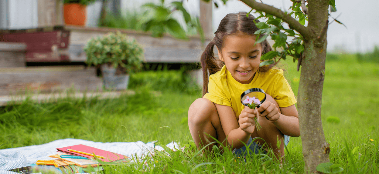 Child with pigtails examining pink flowers with a magnifying glass in grass.