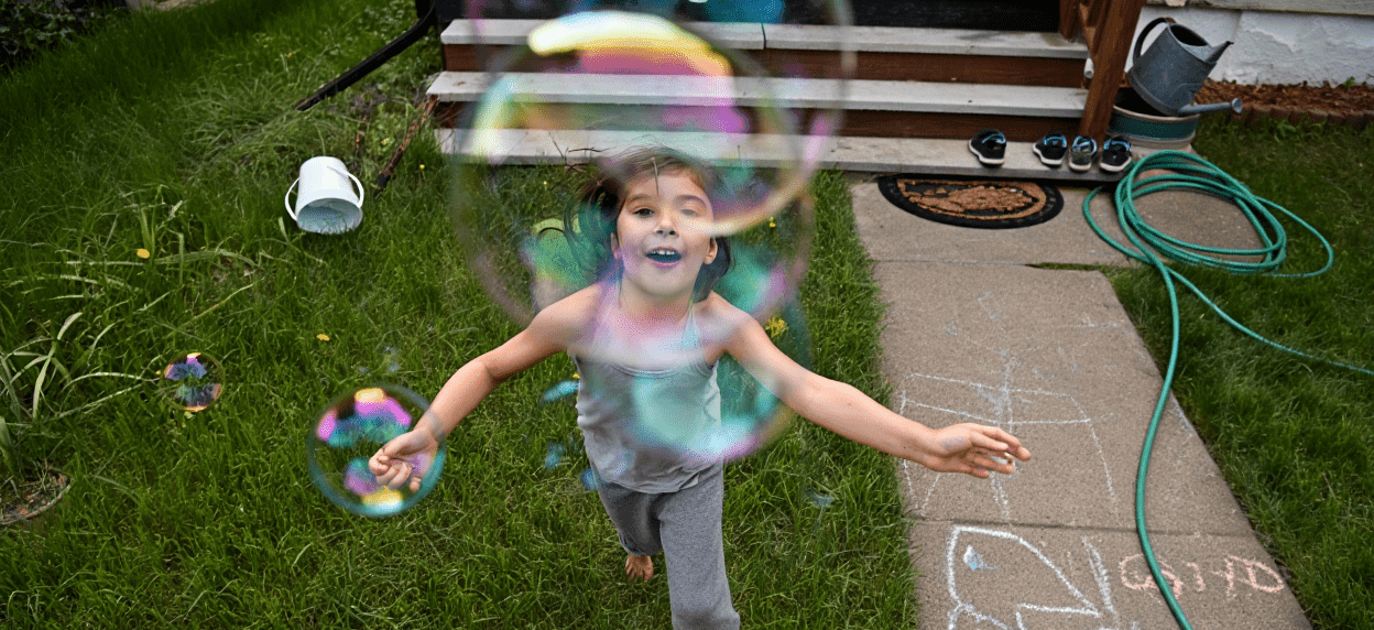 Smiling child playing with colorful bubbles in a green yard.